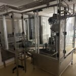 Línea de llenado y taponado de botellas BOC Edwards bajo una cubierta protectora de cristal, con mesas giratorias y cinta transportadora